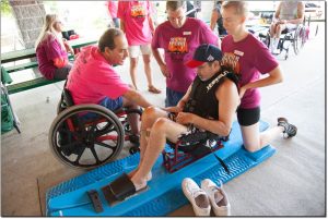 3 adult male volunteers, one in a wheelchair, are helping an adult male skier, who is sitting in a sit ski, in getting fitted for his ski equipment